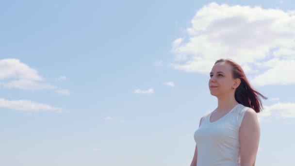 Νεαρή γυναίκα με λευκό φόρεμα στο φόντο του μπλε ουρανού με σύννεφα. Όμορφη γυναίκα χαμογελά, κοιτάζει μακριά και βάζει κομψό καπέλο σε ηλιόλουστο και θυελλώδη καιρό. — Αρχείο Βίντεο