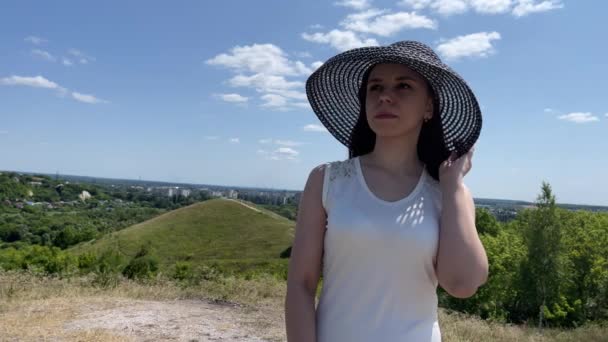Νεαρή Γυναίκα Λευκό Φόρεμα Και Κομψό Καπέλο Στέκεται Λοφώδες Έδαφος — Αρχείο Βίντεο