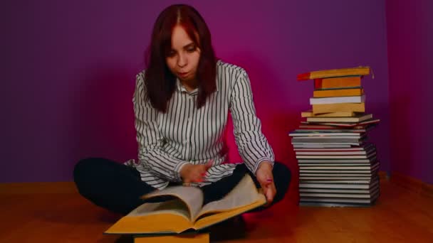 年轻女人坐在地板上 手里拿着一堆书 翻阅着紫色背景的书 穷困潦倒的学生晚上看书 准备上教育课 — 图库视频影像