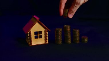 Siyah arka planda madeni para kolonları olan küçük ahşap bir ev. İnsan eli yazı tura atar. Yeni daire ve çatı satın alma kavramı