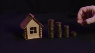 Siyah arka planda madeni para kolonları olan küçük ahşap bir ev. İnsan eli yazı tura atar. Yeni daire ve çatı satın alma kavramı