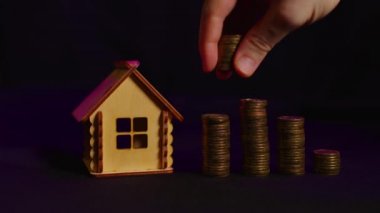 Siyah arka planda madeni para kolonları olan küçük ahşap bir ev. İnsan eli yazı tura atar. Yeni daire ve çatı satın alma kavramı