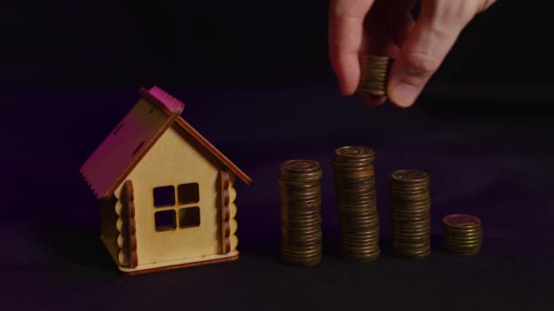 黒い背景にコインの列を持つ小さな木造の家 人間の手はコインを列に入れる 新しいアパートや屋根のオーバーヘッドを購入する概念 — ストック動画