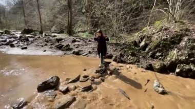 Dişi turist taşların üzerinde nehri dikkatle geçiyor. Genç bir kadın yürüyüşünde engellerin üstesinden gelir.