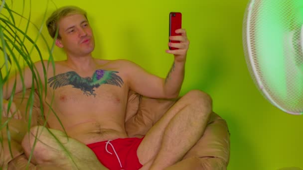 裸の胴を持つ若い男は ファンの前にアームチェアに座って スマートフォンで自撮りを取ります 大人の男はリゾート地として自宅で休暇を楽しんで携帯電話で自分自身の写真を撮る — ストック動画
