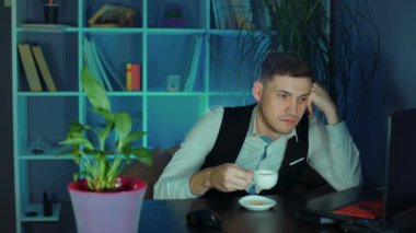 Dizüstü bilgisayarda elinde kahve fincanı tutan uykulu adam. Yorgun ve uykulu bir iş adamı, işyerinde uyukluyor ve kahve içiyor. İş yerinde uyuyan bir adam.