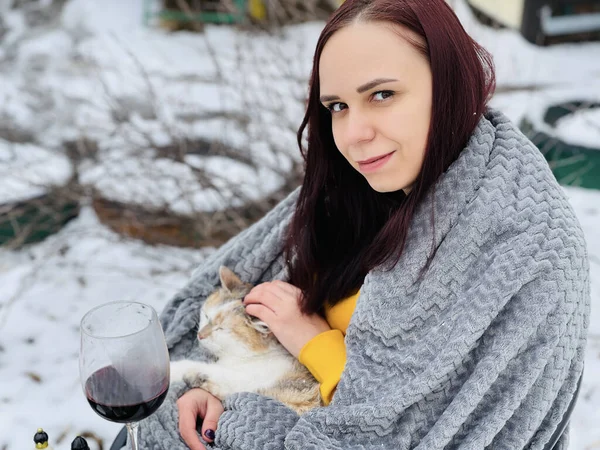 Young woman with domestic cat in hands sitting on street in yard. Female wrapped in grey plaid strokes cute pussycat