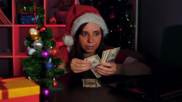 サンタクロースの衣装を着た若い女性は、オフィスのコンピュータの前の椅子に座っている間にお金をカウントします。職場でお金を持って新年のスーツと帽子のビジネス女性マネージャー. — ストック動画