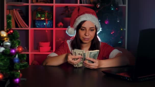 Noel Baba kostümü giymiş genç bir kadın ofiste bir bilgisayarın önünde sandalyede otururken para sayıyor. İş yerinde parası olan takım elbiseli ve şapkalı bir kadın yönetici.. — Stok video