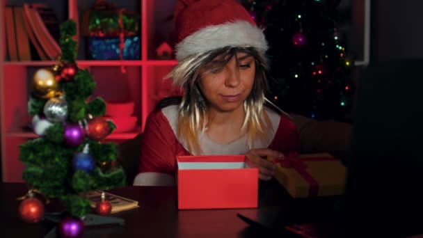 サンタの衣装の若い女性がギフトボックスを開き、それにお金を入れます。新年の雰囲気の中でアームチェアや包装ギフトに座っている従業員。休日を予測する概念. — ストック動画