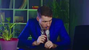 Genç adam ofiste otururken kalemi ısırıyor. Düşünceli iş adamı, modern iş yerinde dizüstü bilgisayarın önünde kendini işe vermiş..