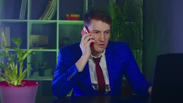 Młody człowiek rozmawia przez telefon komórkowy, siedzi w biurze. Biznesmen przemawia na smartfonie w nowoczesnym miejscu pracy podczas pracy na laptopie. — Wideo stockowe