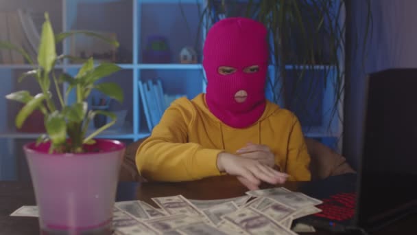 Jonge vrouw in roze bivakmuts verzamelt geld van tafel in het kantoor. Hacker in masker met dollarbiljetten tijdens het zitten in de moderne werkplek. — Stockvideo