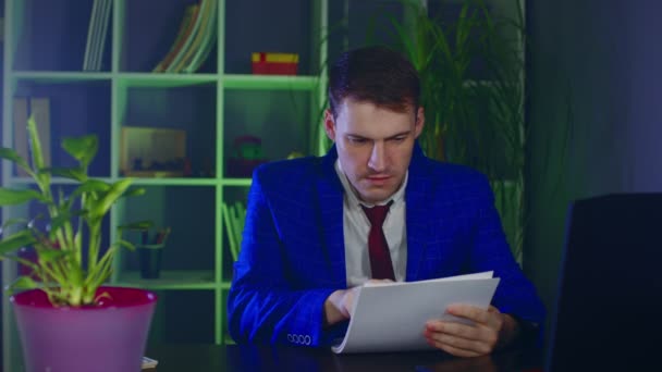 A fiatalember megvizsgálja a dokumentumokat, hivatalban van. Komoly üzletember ellenőrzi a papírokat, ül a laptop előtt a modern munkahelyen. — Stock videók