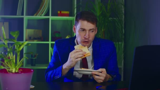 Młody człowiek je kanapki, siedzi w biurze. Biznesmen cieszący się jedzeniem w nowoczesnym miejscu pracy podczas pracy. — Wideo stockowe