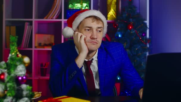 Een jongeman met een kerstmuts werkt op een laptop, zittend op kantoor. Droevige zakenman op moderne werkplek in nieuwjaarssfeer. — Stockvideo