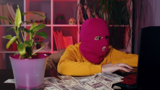 Mujer joven en pasamontañas rosa trabaja en el ordenador portátil en la oficina. Hacker en máscara hace trabajo ilegal mientras está sentado en el lugar de trabajo moderno con dinero en la mesa. — Vídeos de Stock