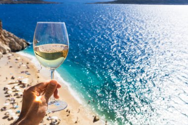 Bir adamın elinde bir bardak beyaz şarap. Deniz kıyısının arka planında şarap ve güneşli bir yaz plajı. Tatil köyünde şarap tatma ve rahatlama.