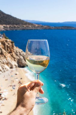 Bir adamın elinde bir bardak beyaz şarap. Deniz kıyısının arka planında şarap ve güneşli bir yaz plajı. Tatil köyünde şarap tatma ve rahatlama.