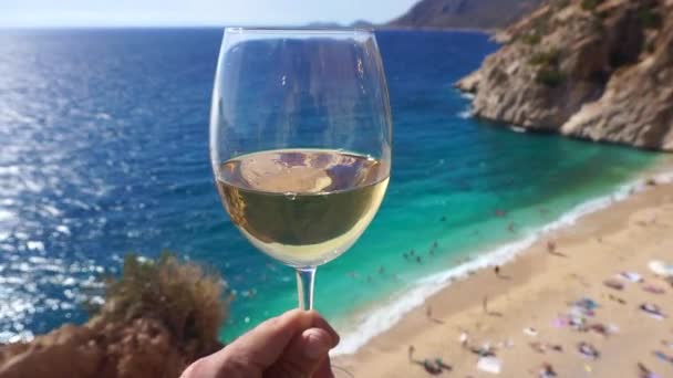 Vino blanco en una copa sobre el telón de fondo de una soleada playa de verano y mar turquesa. Mano con copa de vino y degustación — Vídeos de Stock