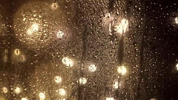 Краплі води течуть по склянці в дощову погоду на розмитому тлі міських вечірніх вулиць і вогнів у боке. Текстура і вплив дощу на вікно — стокове відео