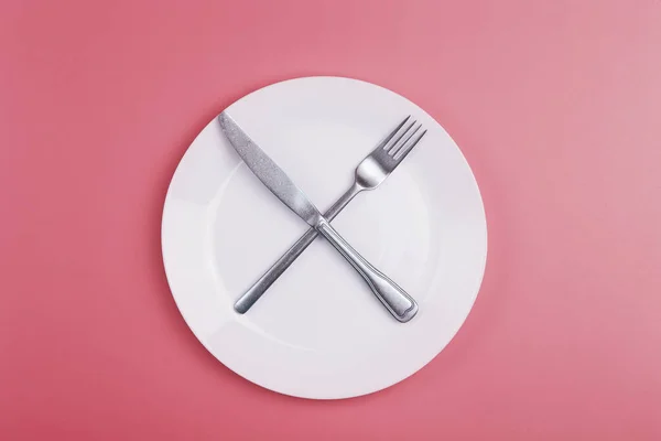 Piastra vuota su sfondo rosa minimale. Piatto vuoto in ceramica bianca con coltello e forchetta sul tavolo dopo aver mangiato. Dieta e concetto di cibo sano. — Foto Stock