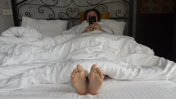 Morning and waking up. Girl in the morning uses a smartphone in bed. Womens feet under the covers in a white bed. Relax, sleep, rest concept. — Stock Video