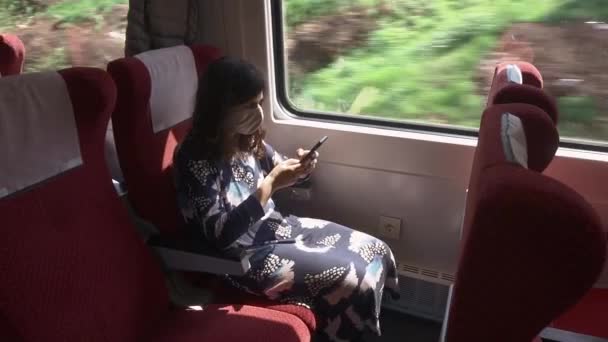 Una niña viaja en tren durante la epidemia de coronavirus COVID-19. Una pasajera con una máscara protectora se sienta en un vagón de tren y usa un teléfono inteligente. Restricción del concepto de circulación y viajes — Vídeo de stock