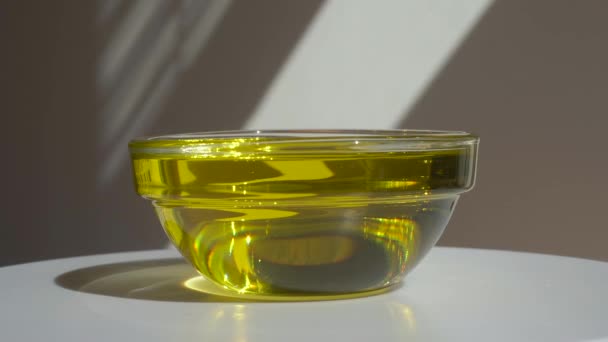 Oil in a bowl on a white background. Yellow organic natural vegetable oil in a glass bowl rotates against the backdrop of a light wall with shadows and sunlight. Cosmetics, food, medicine concept. — Stock Video