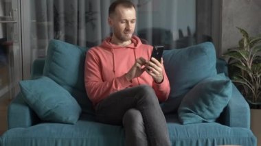 Evindeki koltukta akıllı telefonu olan genç bir Avrupalı adam internet, kurye ve online alışveriş kullanıyor. Uygulamalar, iletişim ve web sörfü kavramı