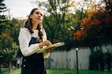 Bir yaz parkında kalemlerle resim yapan genç bir kızın portresi. Sakinlik, ilham ve yaratıcılık atmosferi doğayla çevrili. Yumuşak doğal ışık, arka planda yeşillik, ve bir an