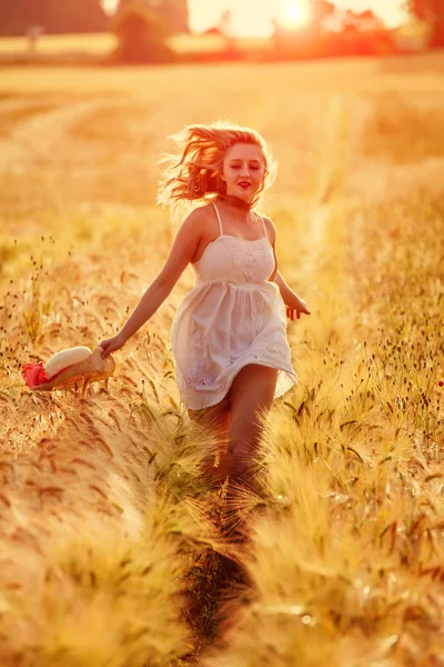 Happy young blonde girl in white dress with straw hat running th