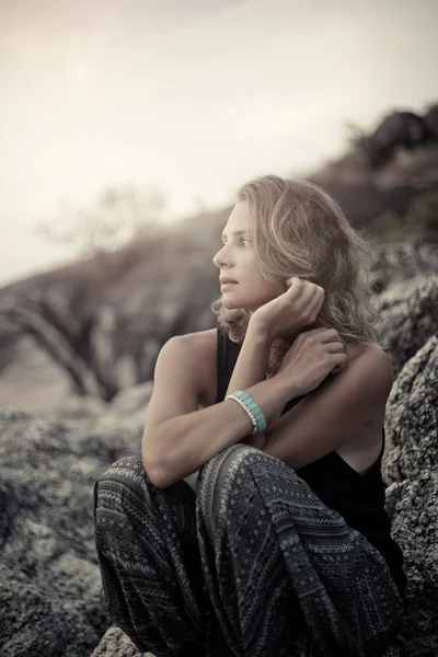Hermosa joven sentada en las rocas al atardecer fondo — Foto de Stock