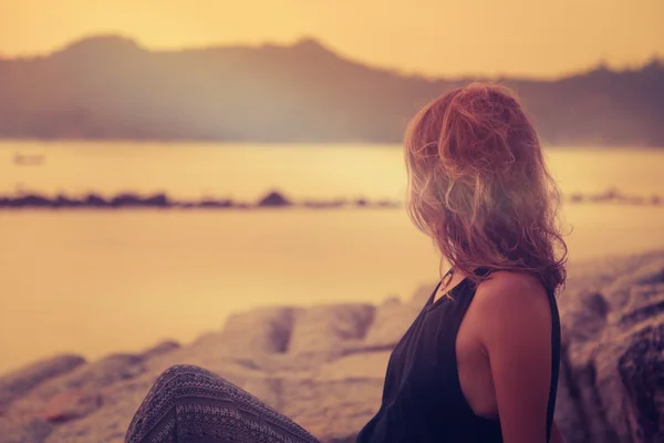 Ung kvinna sitter på klipporna vid havet och tittar på solen — Stockfoto