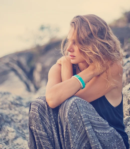 Hermosa joven sentada en las rocas al atardecer fondo — Foto de Stock