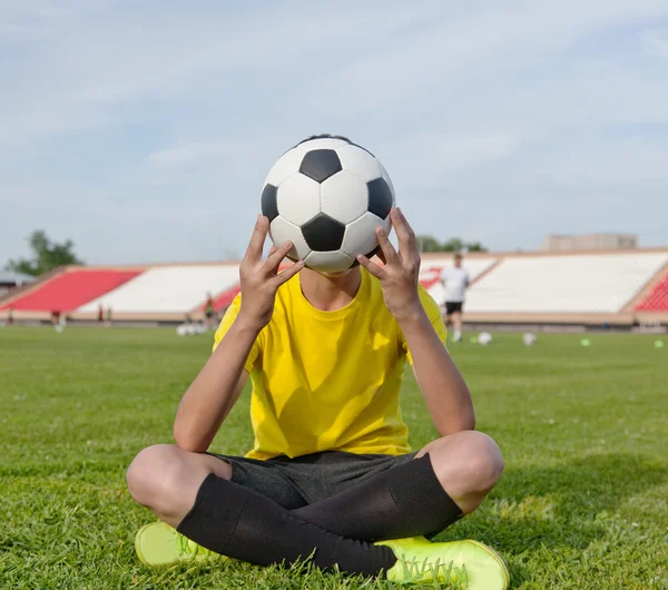 Chłopiec siedzi na trawie w stadion piłkarski i posiada socc — Zdjęcie stockowe