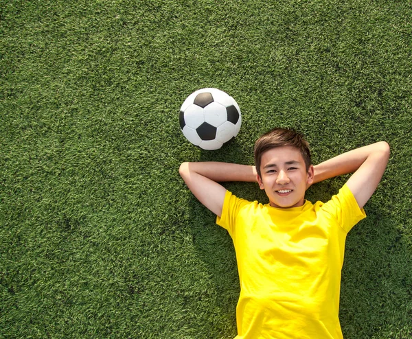 Щасливий хлопчик-підліток з футбольним м'ячем, що лежить на зеленому газоні — стокове фото