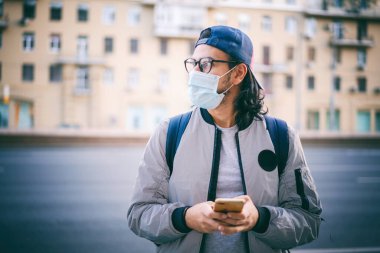 Gözlüklü, koruyucu maskeli, sırt çantalı ve beysbol şapkalı sevimli esmer hippi, akıllı telefonlu, yeni normal yaşam tarzı salgını olan adam.