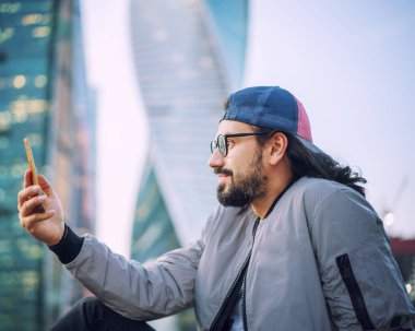 Gözlüklü şık esmer hippi, büyük şehirdeki akıllı telefon ekranına, rahat bir hayat için mobil uygulamalara ve mutlu insanlara bakıyor.