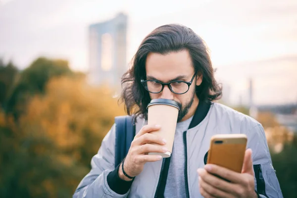 メガネのかわいいスタイリッシュなブルネットのヒップスターは 大都市で携帯電話のスマートフォン画面を見て コーヒーを飲む — ストック写真