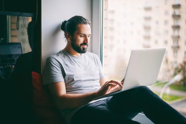 魅力的な若いですダークヘアの男は自宅でノートパソコンで作業 街の景色と窓辺に座って — ストック写真
