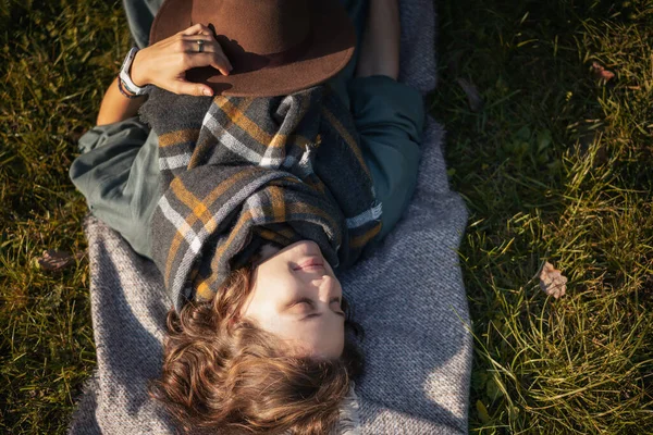 帽子の秋の公園で目を閉じた美しい巻き毛の若い女性と草の上に横たわる有料スカーフ — ストック写真