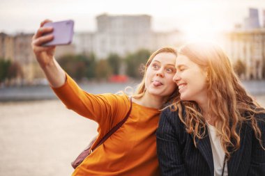 İki güzel genç bayan arkadaş şehirde birlikte yürürken, akıllı telefon kullanarak selfie çekiyorlar.