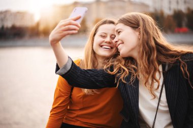 İki güzel genç bayan arkadaş şehirde birlikte yürürken, akıllı telefon kullanarak selfie çekiyorlar.