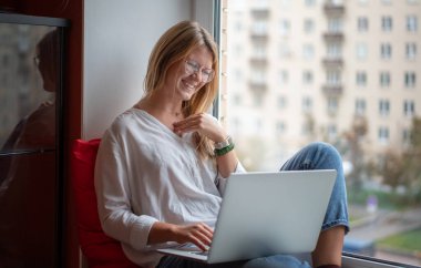 Beyaz tişörtlü, uzun kızıl saçlı genç bir kadın evdeki dizüstü bilgisayarda çalışıyor, uzaktan kumandalı serbest çalışan. Pencere kenarında otururken