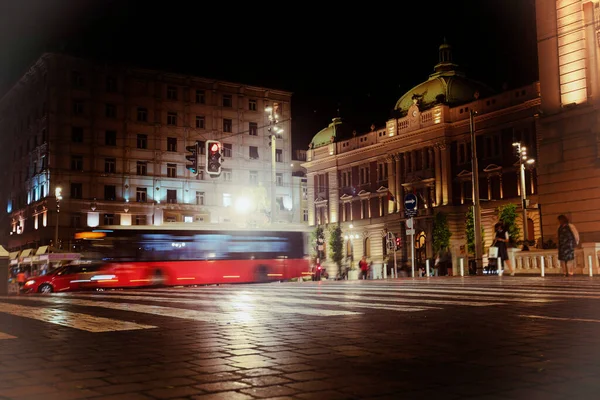Pedestrian crossing in the historic center of Belgrade, red bus on the road and blurred figures of people in the night
