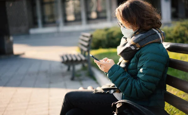 Piękna młoda Europejka w masce ochronnej na ulicy miejskiej ze smartfonem w rękach siedzi na ławce — Zdjęcie stockowe