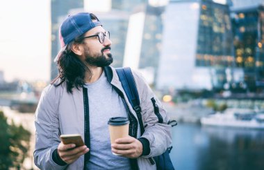 Gözlüklü sevimli esmer hippi büyük bir şehirde cep telefonunun ekranına bakar ve kahve içer.