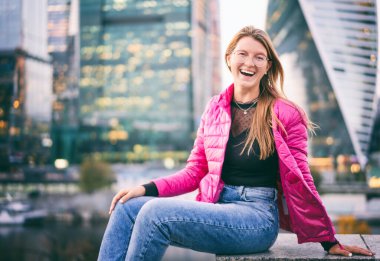 Genç, mutlu, gözlüklü, uzun kızıl saçlı, parlak pembe ceketli güzel bir kadın. Büyük şehirde hayatın tadını çıkarıyor.