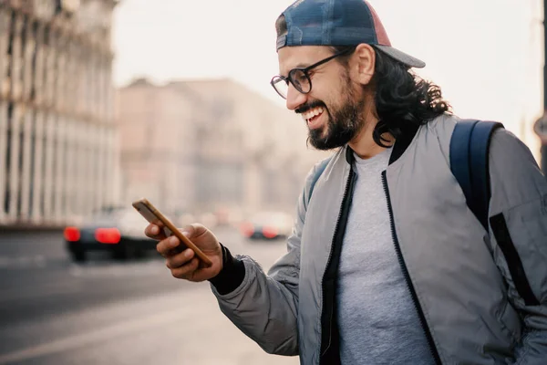 かわいい幸せな笑顔ブルネットヒップスターバックパック付き 大都市で彼の手の中にスマートフォンを持つ男 — ストック写真